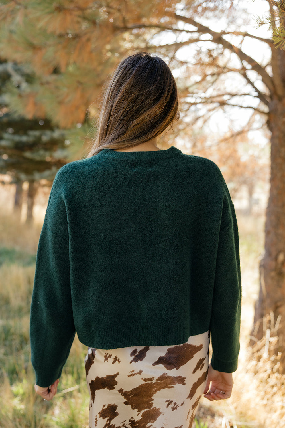 Person wearing a green sweater and patterned skirt standing in a forest.
