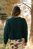 Person wearing a green sweater and patterned skirt standing in a forest.