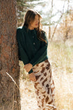 Woman leaning against a tree in a natural setting with dry grass and trees.