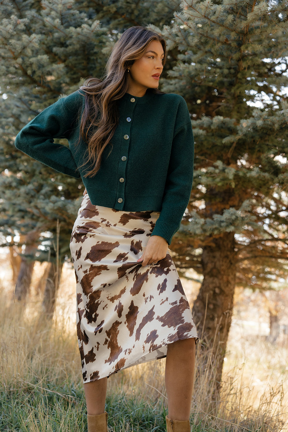 Woman wearing a green cardigan and cow print skirt standing in a forested area.