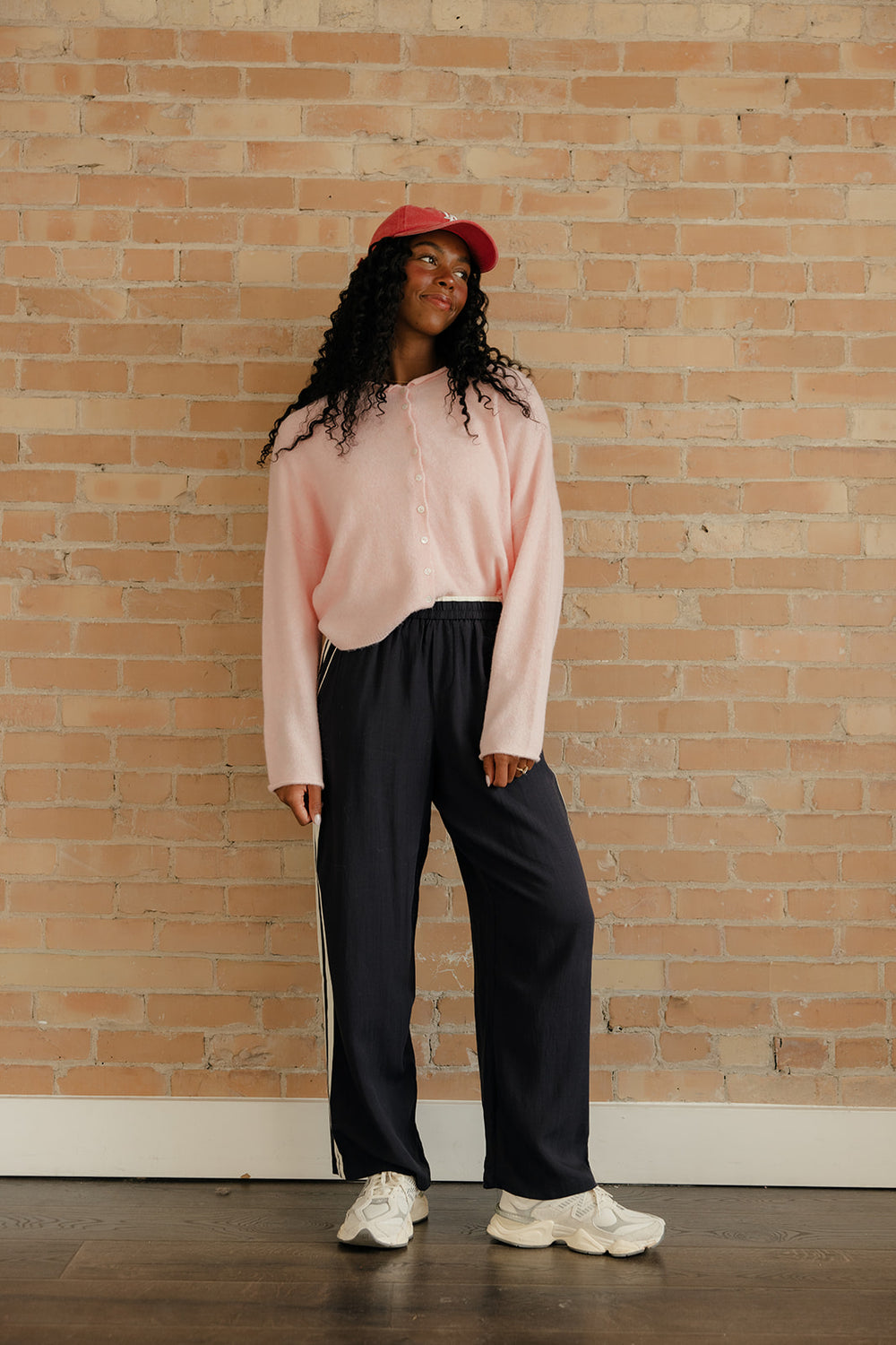 Person wearing a pink sweater and black pants standing against a brick wall.