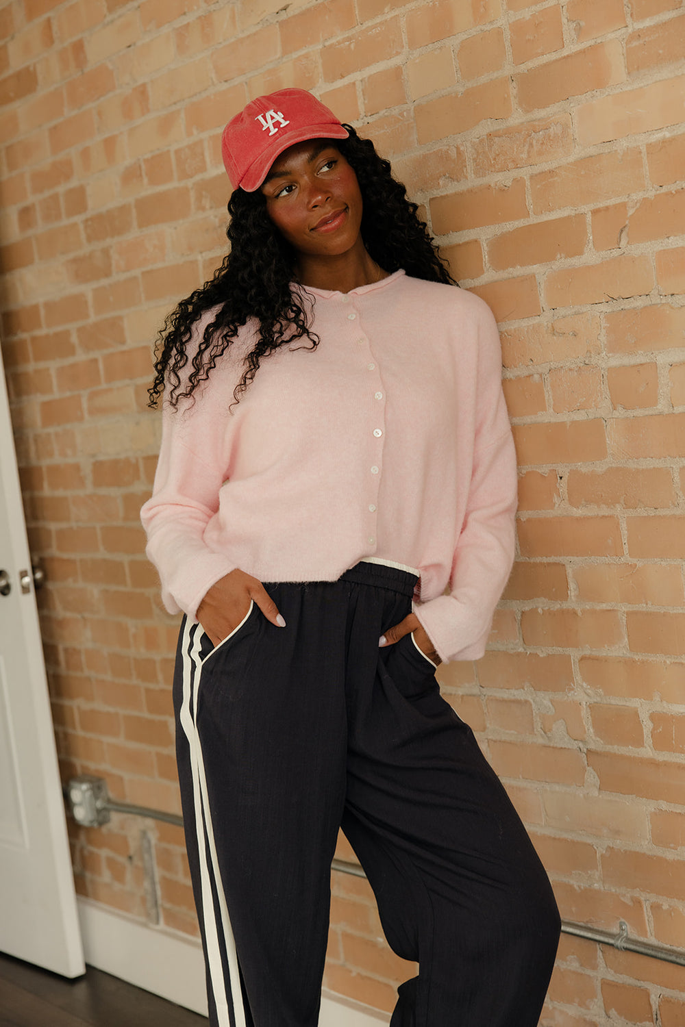 Person wearing a pink sweater and black pants with white stripes, standing against a brick wall.