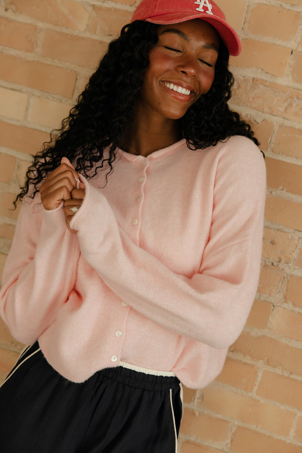 Woman wearing a pink cardigan and red cap against a brick wall