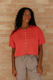 Woman wearing a red cropped top with buttons and checkered pants against a brick wall.