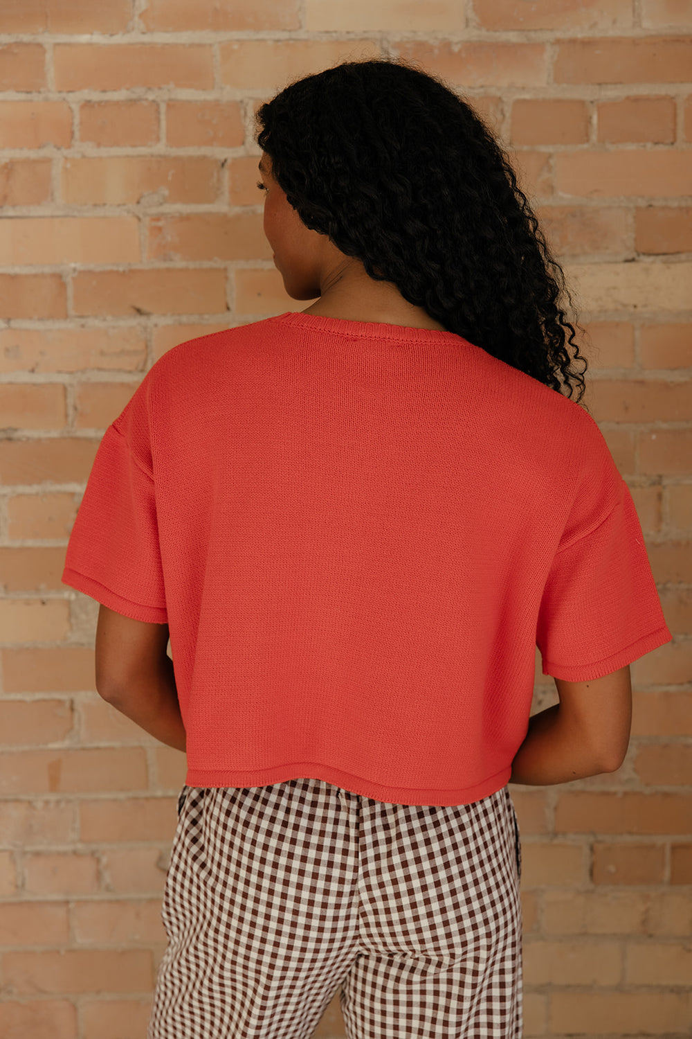 Person wearing a red t-shirt and checkered pants against a brick wall.