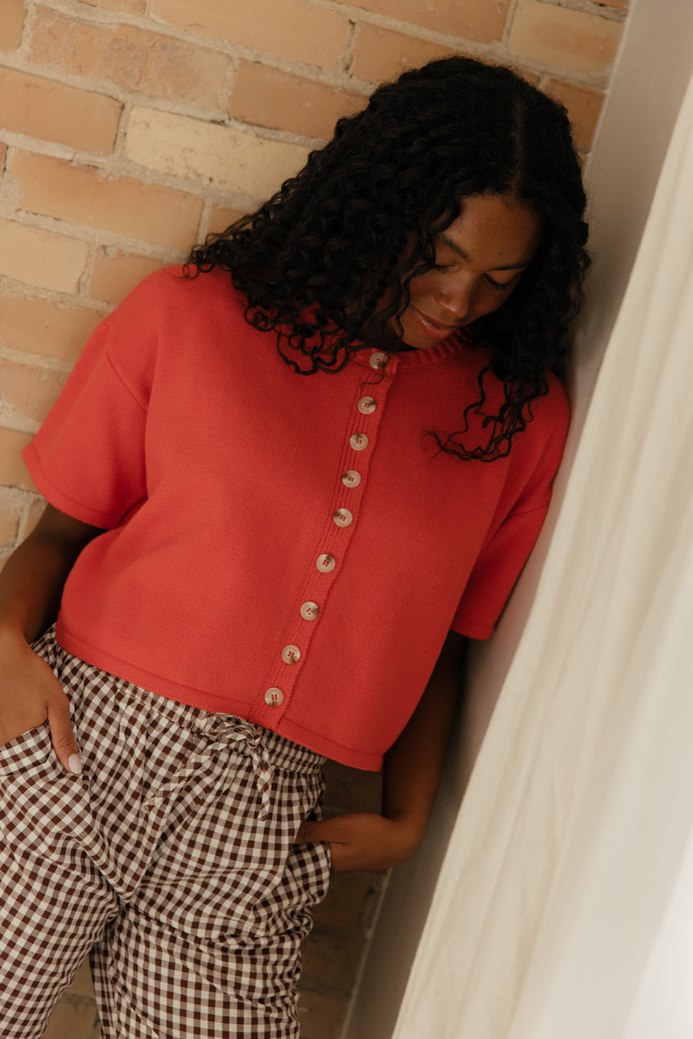Person wearing a red shirt with button details and checkered pants against a brick wall.