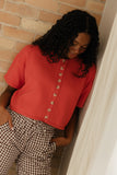Person wearing a red shirt with button details and checkered pants against a brick wall.