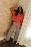Woman in a red blouse and checkered pants standing against a brick wall.