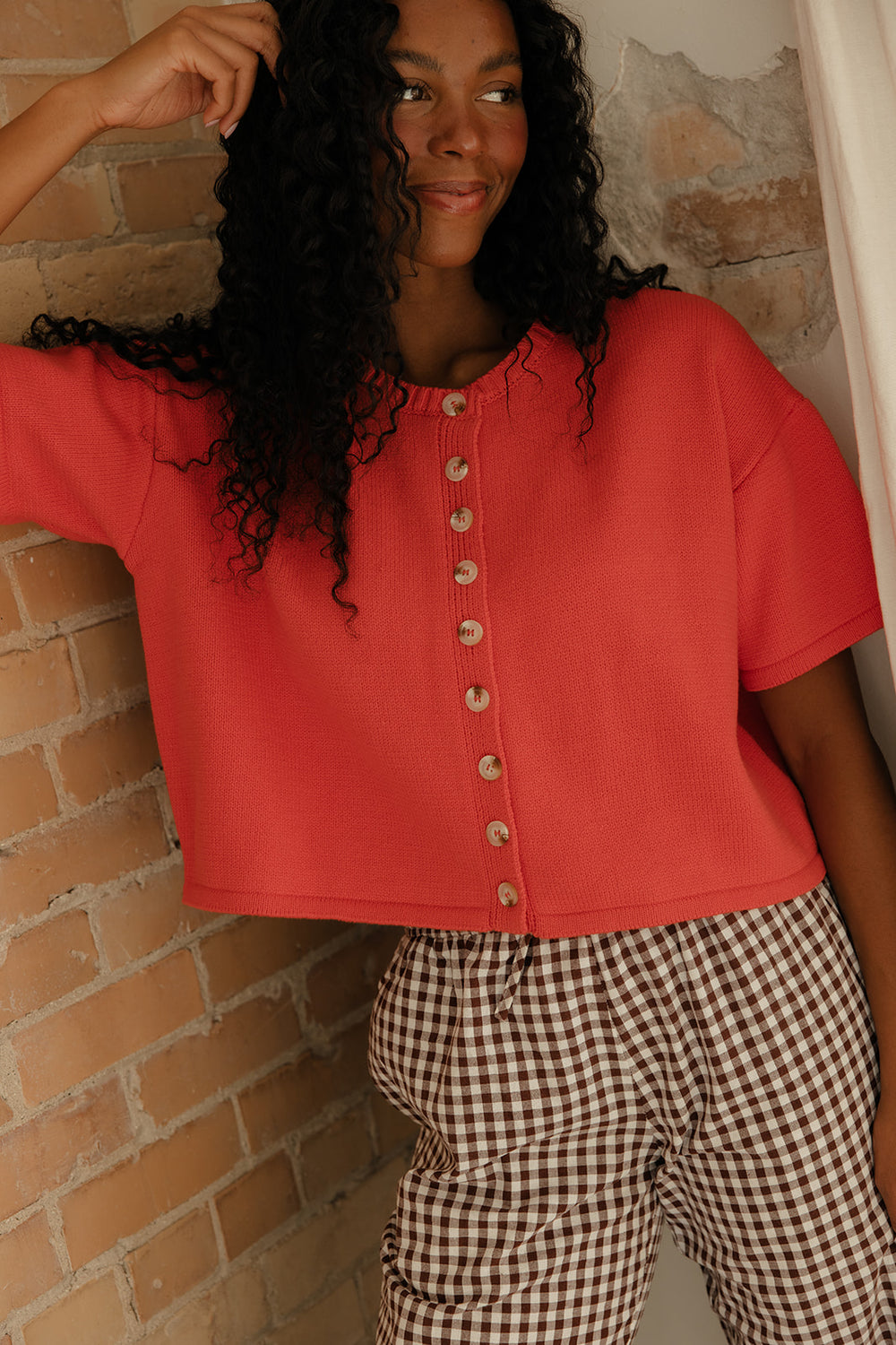 Person wearing a red button-up shirt and checkered pants against a brick wall.