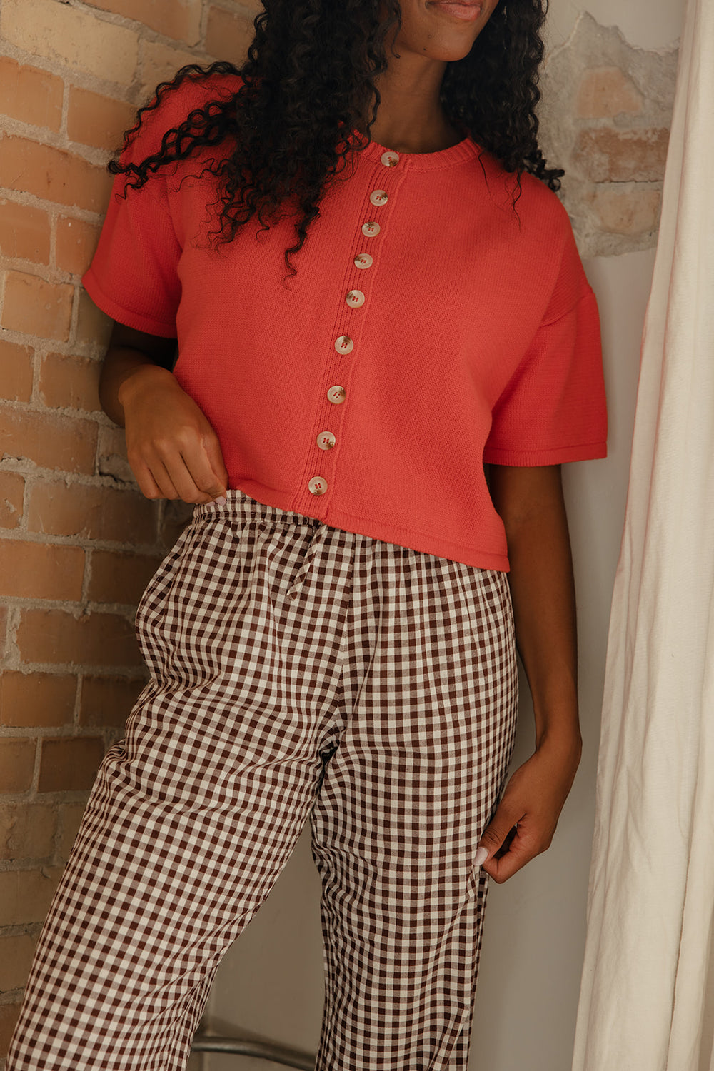 Person wearing a red top with buttons and checkered pants against a brick wall.