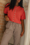 Person wearing a red top with buttons and checkered pants against a brick wall.