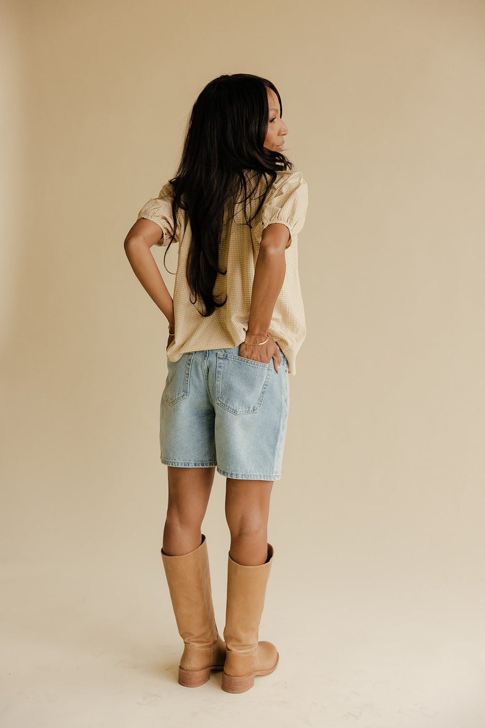 Woman wearing a beige shirt, light blue shorts, and tan boots against a beige background