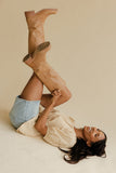 Woman lying on a beige surface with one leg raised wearing tan knee-high boots.