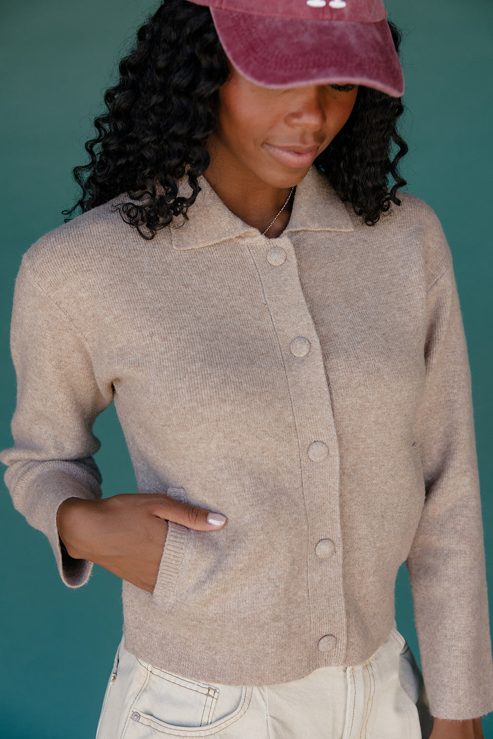 Woman wearing a beige cardigan and pink cap against a teal background