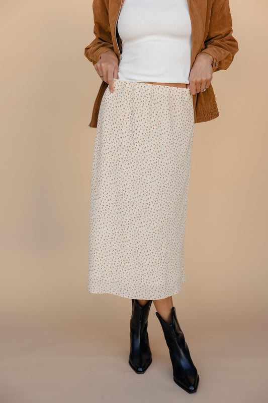 Person wearing a patterned skirt, white top, brown cardigan, and black boots on a beige background
