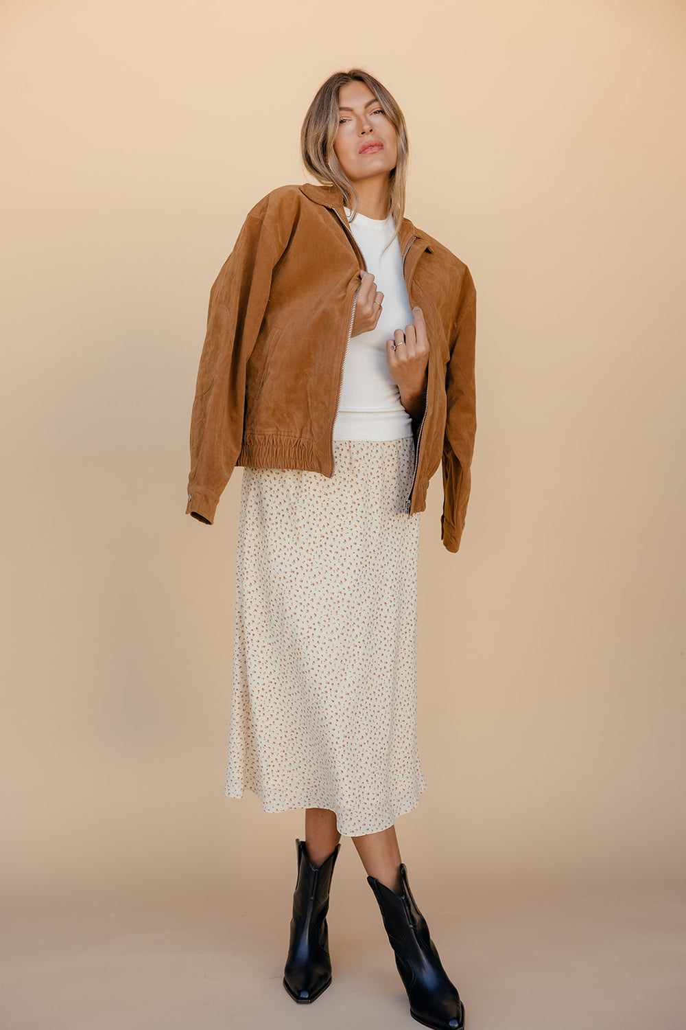 Woman wearing a brown jacket, white top, and patterned skirt against a beige background