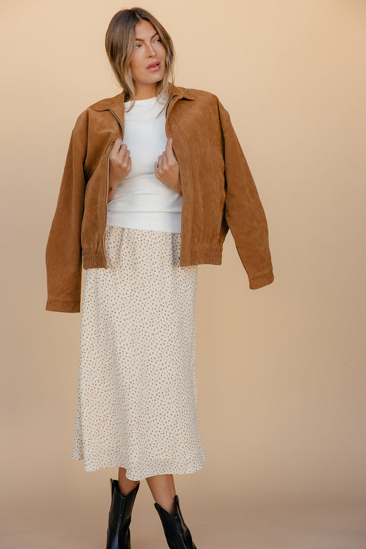 Woman wearing a brown jacket, white top, and patterned skirt against a beige background