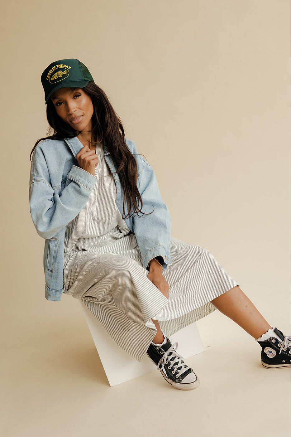Woman sitting on a white stool wearing a denim jacket, white dress, and black sneakers on a beige background
