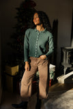 Woman in a green sweater and brown pants standing in a room with a Christmas tree.