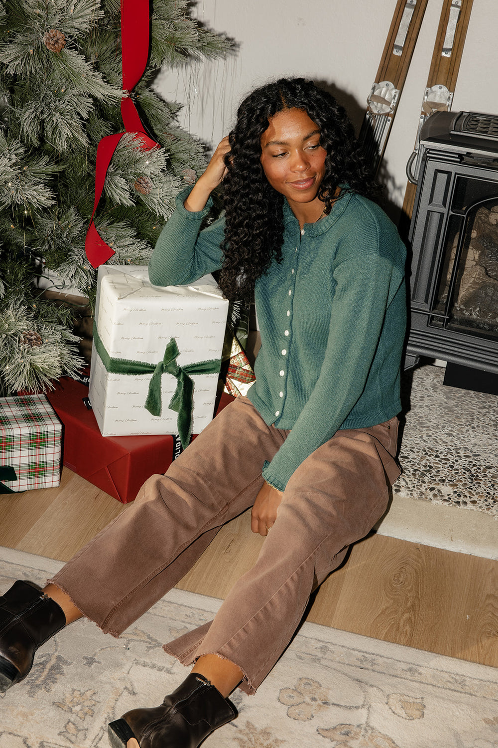 Woman sitting by a Christmas tree with presents, wearing a green shirt and brown pants.