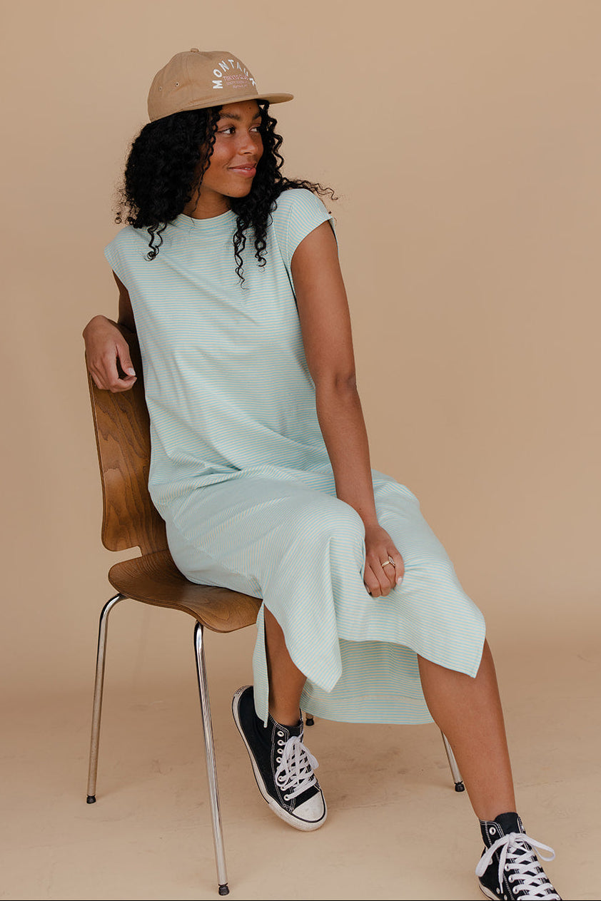 Woman sitting on a chair wearing a light green dress, black sneakers, and a beige hat against a beige background.