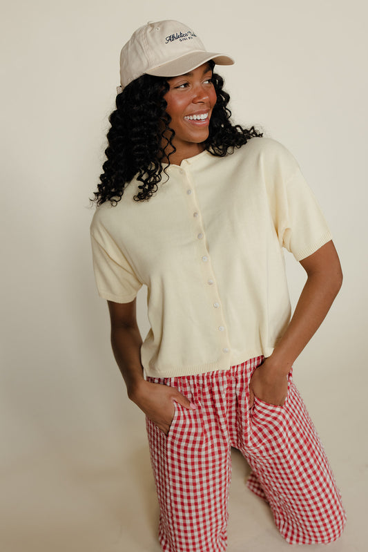 Woman wearing a cream shirt and red checkered shorts with a beige cap on a plain background
