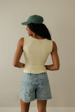 Person wearing a beige sleeveless top, denim shorts, and a green cap against a plain background