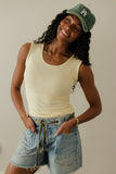 Woman wearing a yellow tank top, denim shorts, and a green cap with a logo on a plain background
