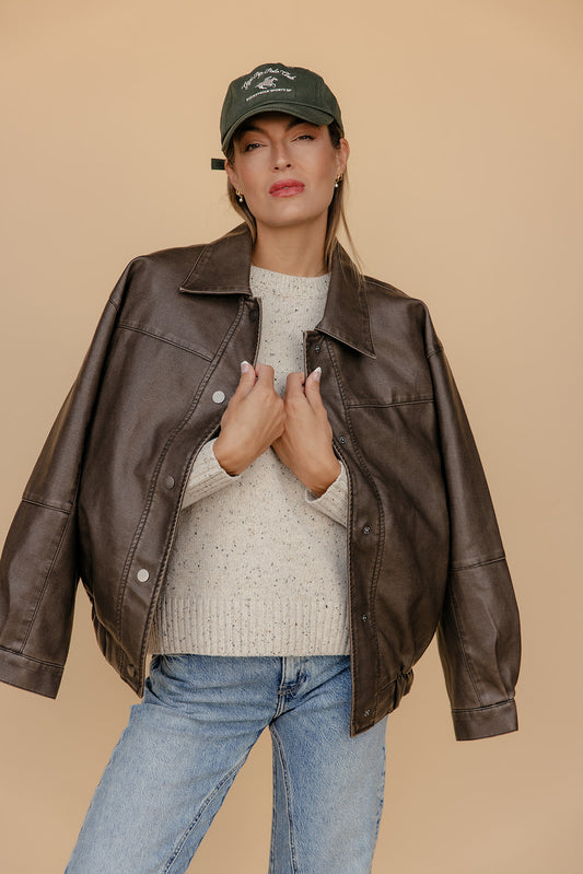 Woman wearing a brown leather jacket, beige sweater, blue jeans, and a green cap on a beige background