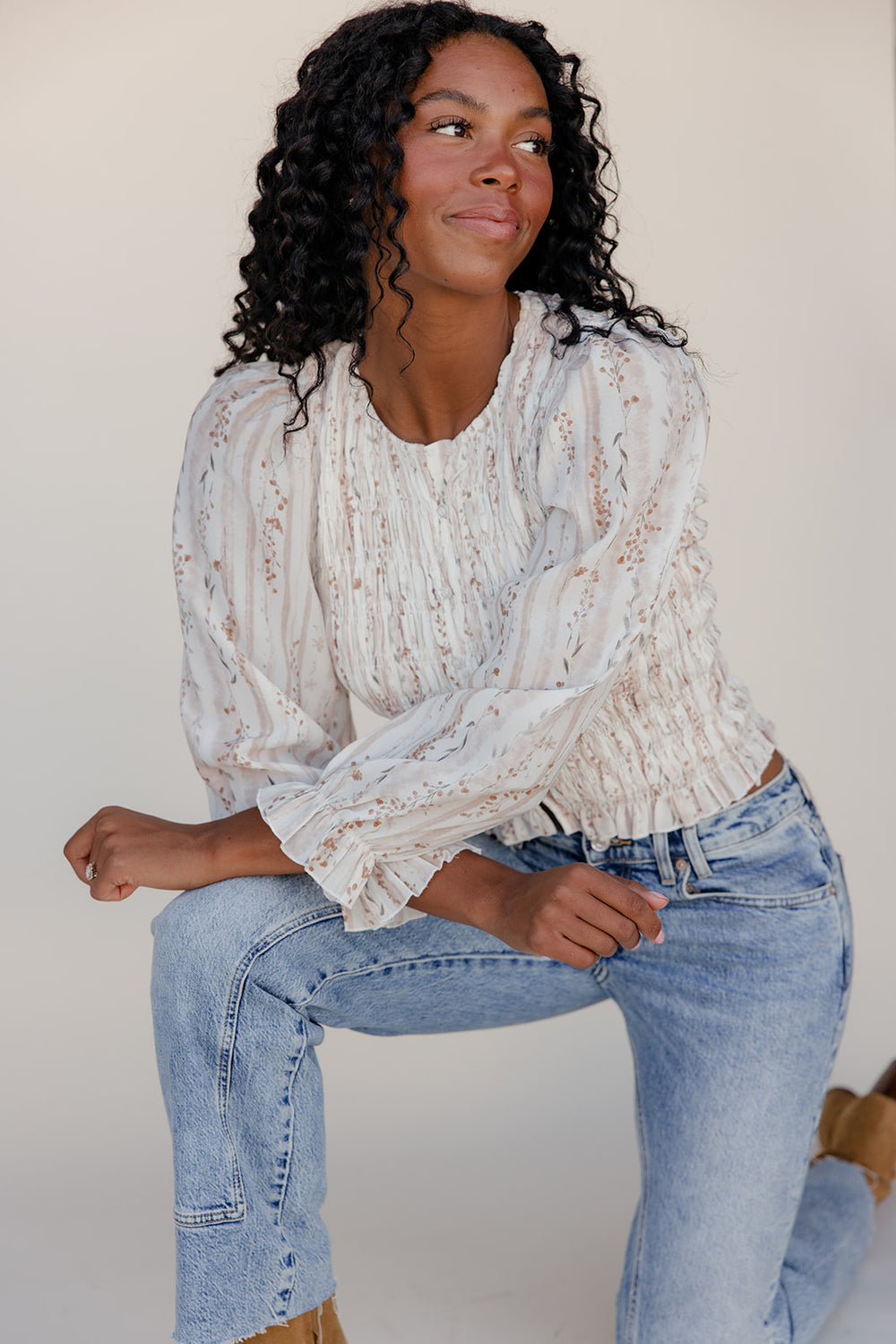 Woman wearing a white lace top and blue jeans sitting against a plain background