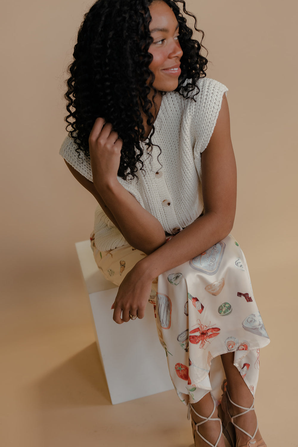 Woman sitting on a white stool wearing a patterned top and floral skirt against a beige background