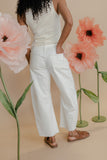 Person wearing a white sleeveless top and pants standing among large pink flowers on a beige background