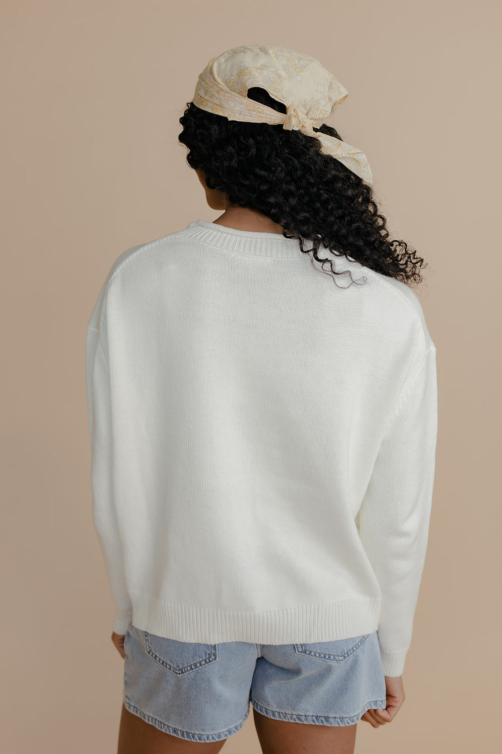 Person wearing a white sweater and denim shorts with a beige headscarf on a beige background