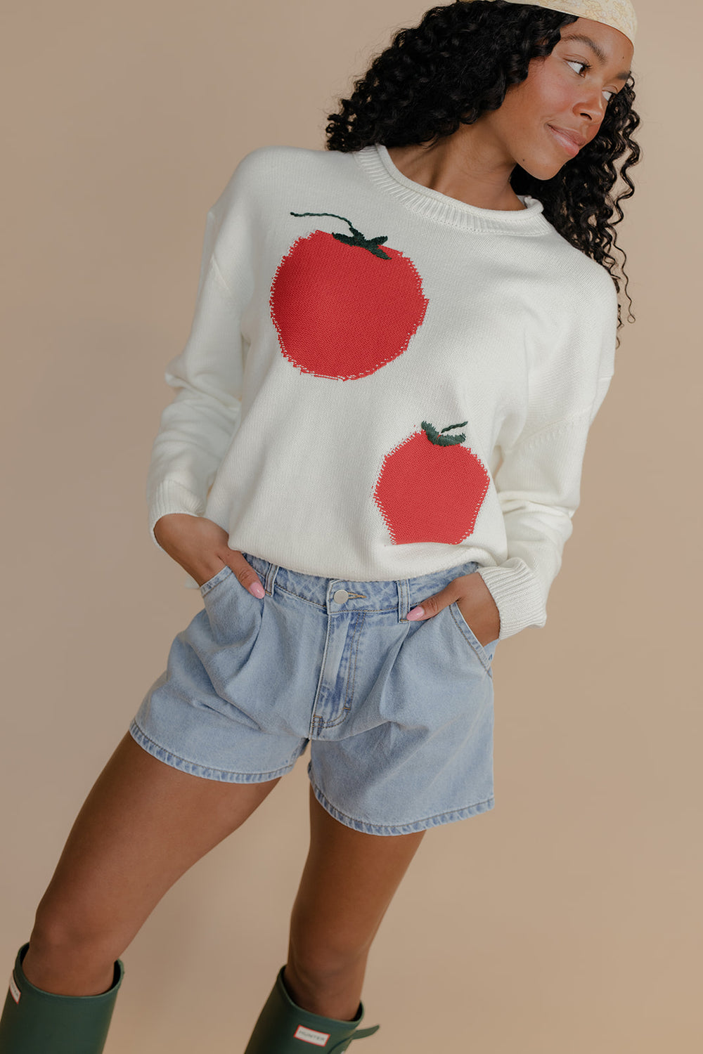 Woman wearing a white sweater with red apple designs and denim shorts on a beige background