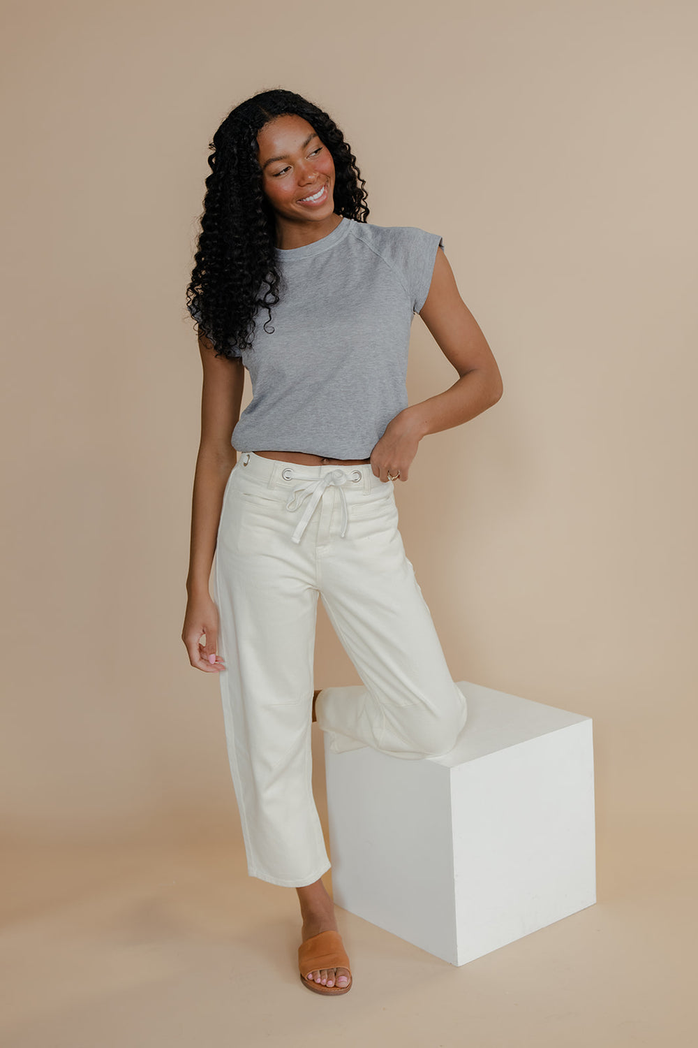 Woman wearing a gray sleeveless top and white pants standing on a white block against a beige background