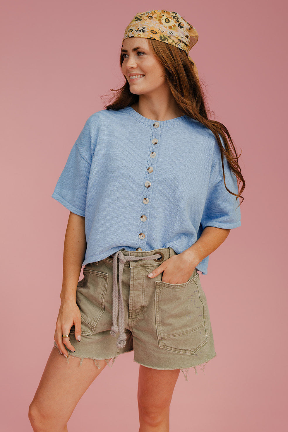 Woman wearing a blue shirt and green shorts with a floral headband on a pink background