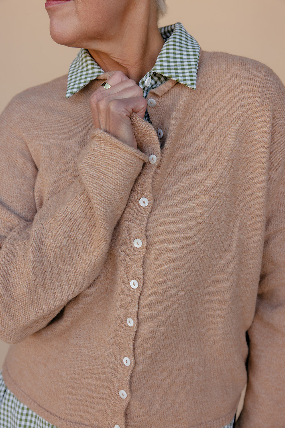 Close-up of a person wearing a brown cardigan with a checkered collar against a beige background