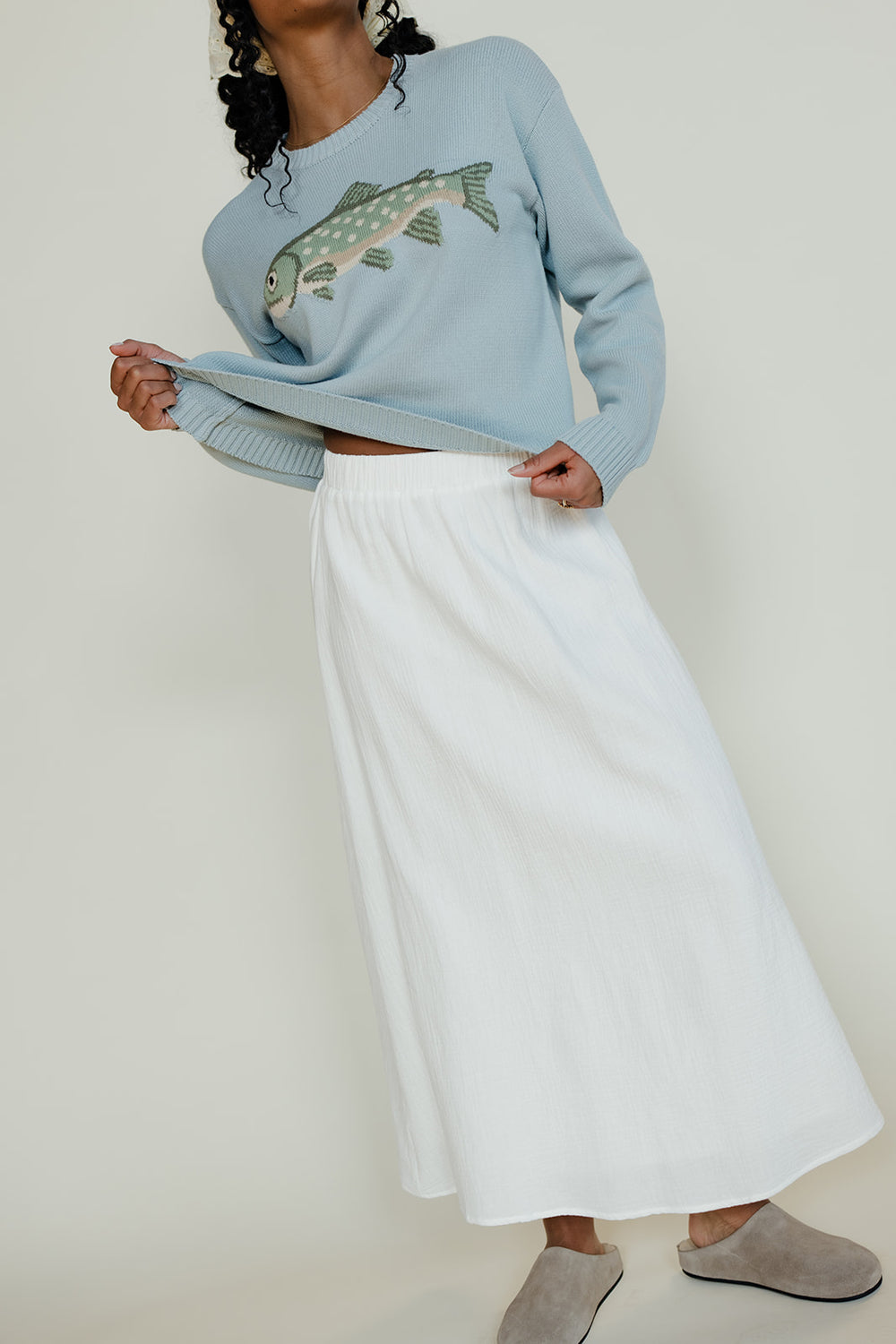 Person wearing a light blue sweater with a fish design and white pants on a plain background