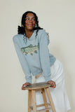 Woman wearing a light blue sweater with a fish design, sitting on a wooden stool against a plain background.