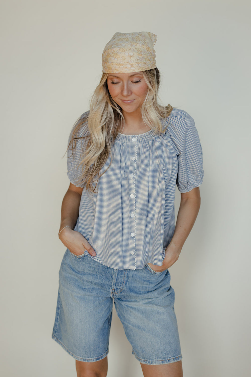 Woman wearing a blue checkered blouse and denim shorts with a beige headscarf against a plain background
