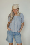 Woman wearing a blue checkered blouse and denim shorts with a beige headscarf against a plain background
