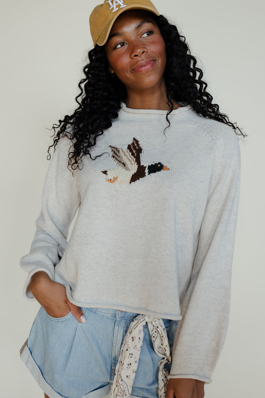 Woman wearing a light gray sweatshirt with a graphic design, denim shorts, and a beige cap.
