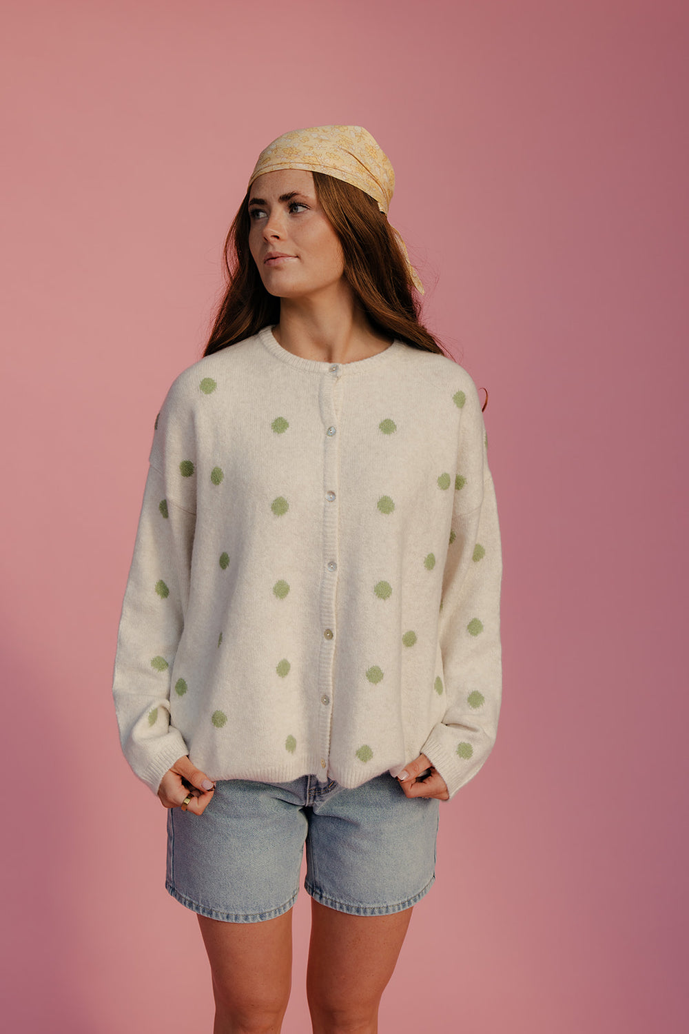 Woman wearing a light-colored cardigan with green polka dots, denim shorts, and a beige headband against a pink background.