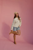 Woman posing in a polka dot sweater, denim shorts, and brown boots against a pink background
