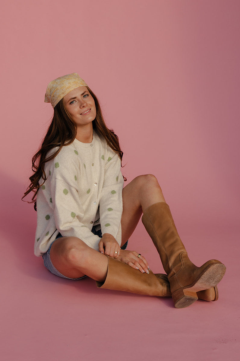 Woman sitting on a pink background wearing a white patterned sweater, denim shorts, and brown knee-high boots.