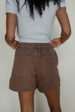 Person wearing brown shorts and a light gray top against a plain background