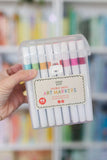 Pack of double-ended art markers held by a hand with a blurred bookshelf background