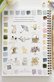 Art book with watercolor illustrations on a white surface with a plant and paint bottle.