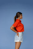 Woman wearing an orange top and denim shorts against a blue background