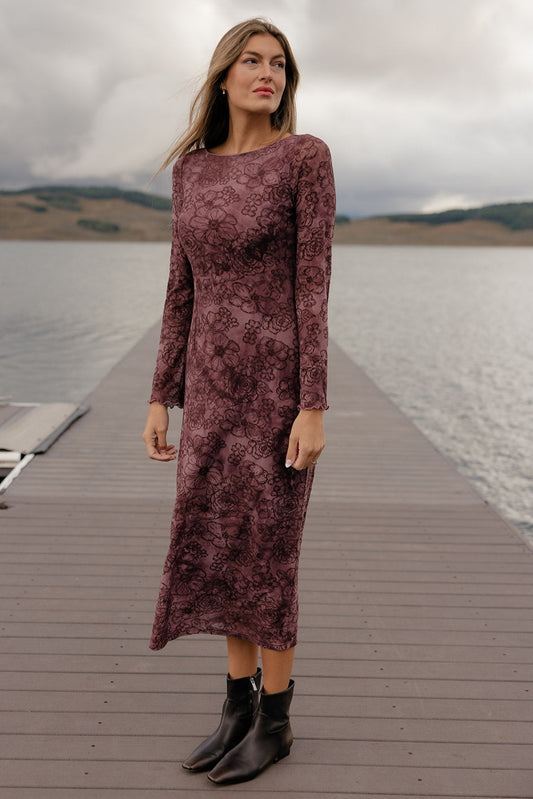Woman in a long, burgundy dress standing on a wooden dock by a lake with mountains in the background.